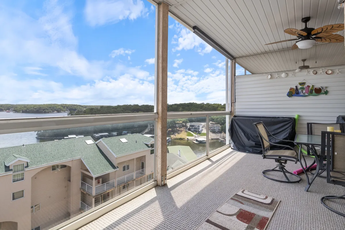 A scenic balcony view with a glass railing overlooking the water, an outdoor dining table and chairs, and tasteful decor. The tranquil setting is ideal for al fresco dining, with a ceiling fan and wall art adding comfort and charm.