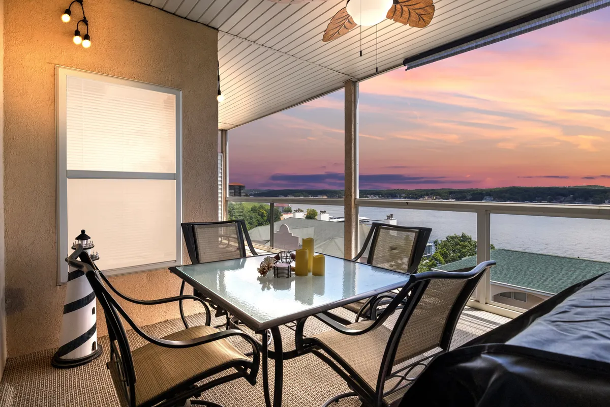 The image showcases a serene waterfront patio at sunset, featuring a glass dining table surrounded by plush chairs, a stylish hanging light fixture, and a decorative lighthouse accent. The patio overlooks the stunning, open expanse of Lake of the Ozarks' main channel, inviting guests to relax, dine, and soak in breathtaking, unobstructed water views.