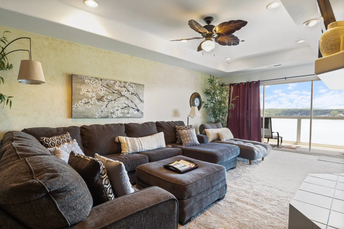 This image shows a spacious living room with a large sectional sofa facing floor-to-ceiling windows that offer stunning views of the lake beyond. The room is filled with natural light and includes tasteful artwork, decorative plants, and a cozy, welcoming atmosphere perfect for group gatherings and relaxing.