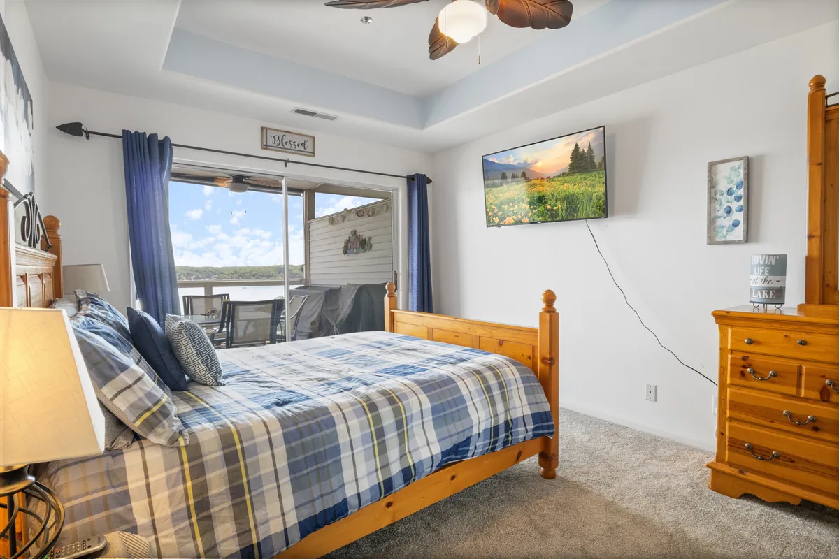 A comfortable bedroom featuring a plaid bed, large window with expansive views, a wall-mounted TV, and wooden furnishings. The room feels cozy and inviting, with natural light flooding the space and offering a peek at the serene outdoors.