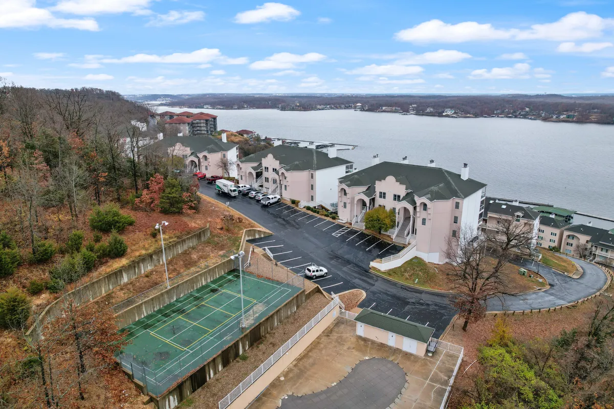 This exterior image captures the lakefront setting with a visible tennis court, adjacent buildings, and a parking area. The calm waters, open space, and recreational facilities evoke the sense of relaxation and group activities that are central to the condo's appeal.
