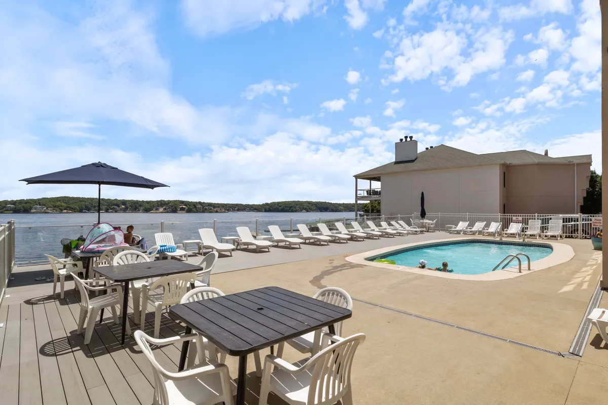 A sparkling swimming pool surrounded by lounge chairs and dining tables, all oriented toward a gorgeous lake view. The ambiance is leisurely, with the option to sunbathe, swim, or socialize by the water's edge.