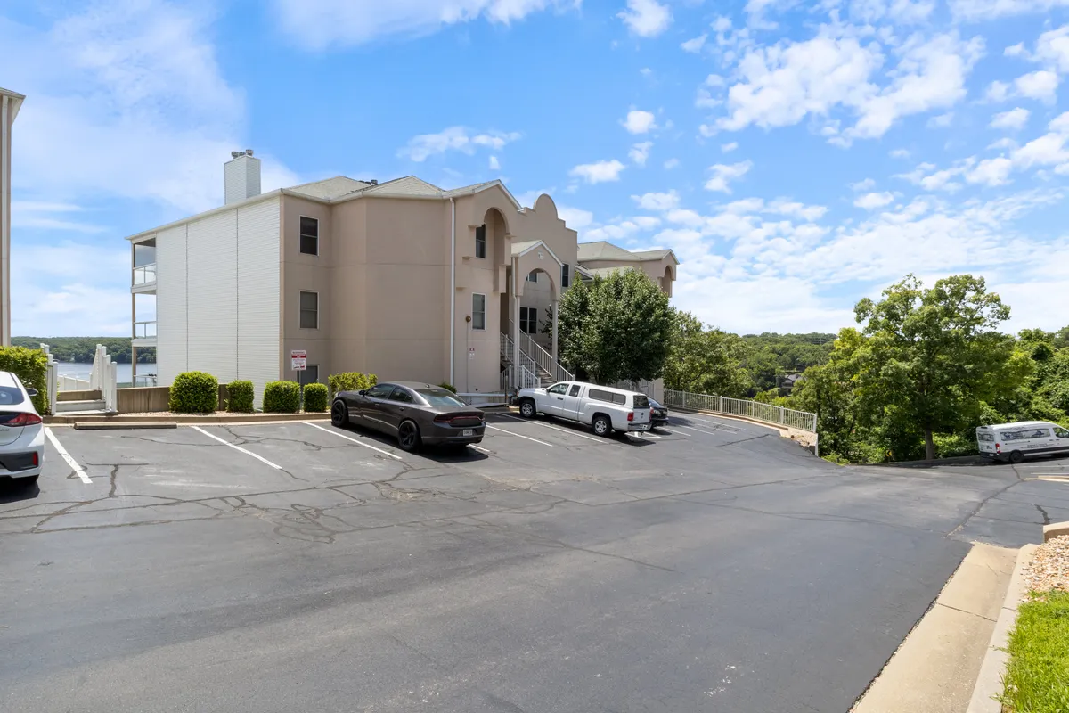 An exterior view of a multi-unit building surrounded by greenery with an ample parking area. The scene is inviting, with space for casual gatherings and easy arrivals and departures, set against a backdrop of nature.
