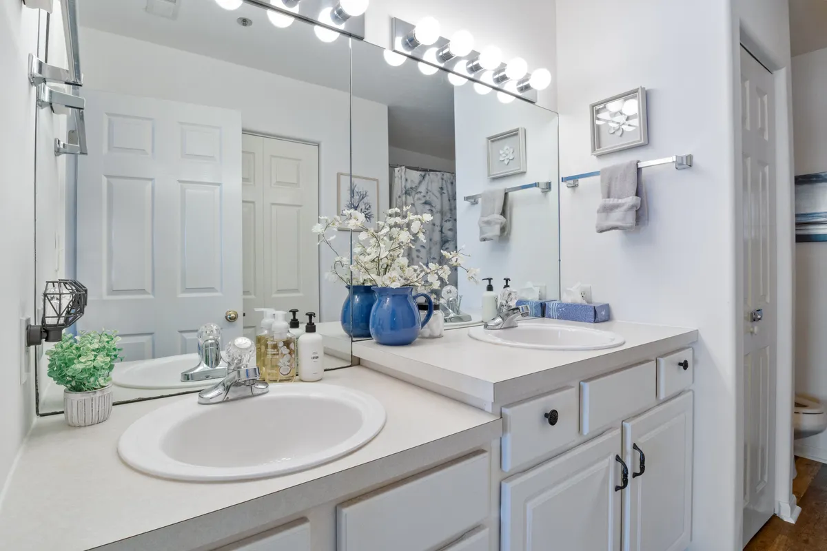 A modern bathroom featuring double sinks, a large mirror, blue floral accents, and soothing wall art. The space is bright, fresh, and designed for both practicality and relaxation, echoing the spa-like comfort of the baths described.