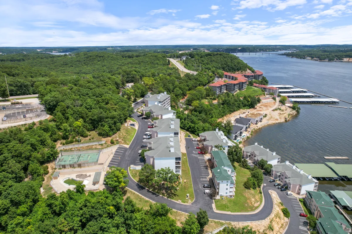 This image shows a waterfront resort property with a well-maintained marina and surrounding greenery. The marina features multiple boat slips right outside the condo buildings, with boats docked and the lake stretching into the distance. The natural landscape and roadways suggest easy access for outdoor activities and exploration.